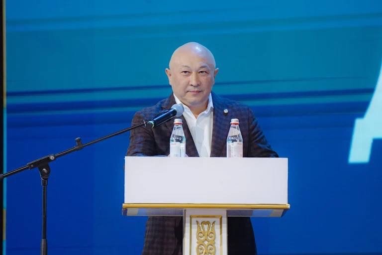 A man in business attire speaking at a podium with a blue background and a partial view of the Qazaqstan Football League logo.